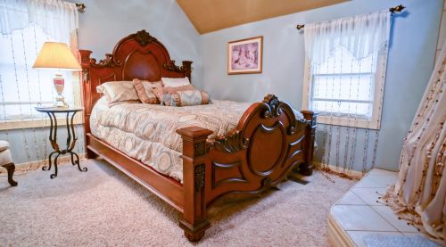 Cozy bedroom featuring a wooden bed, patterned bedding, a lamp, and light blue walls with sheer curtains.