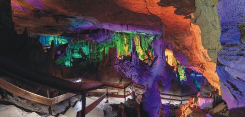 A colorful cave interior with illuminated stalactites and stalagmites, featuring wooden walkways and vibrant lighting.