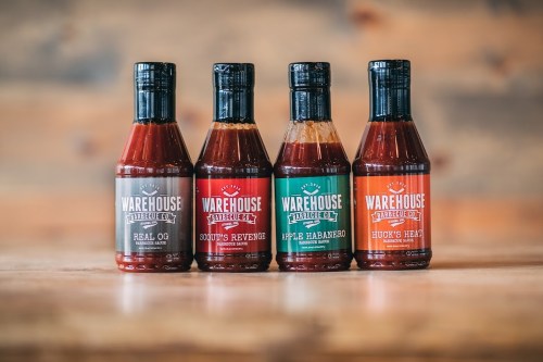 Four bottles of Warehouse barbecue sauce in various flavors, arranged on a wooden surface with a rustic background.