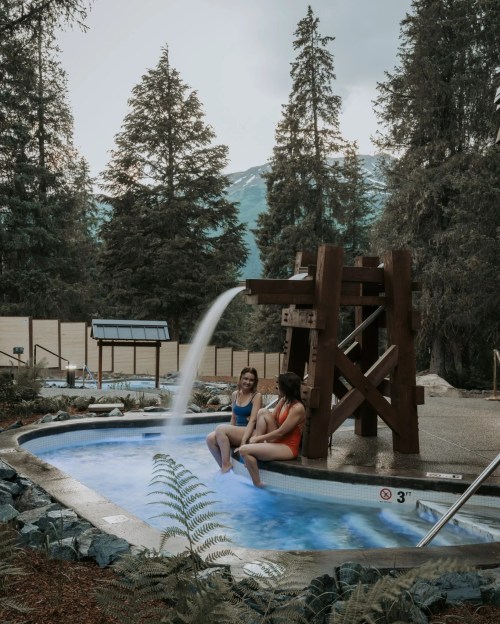 Two women relax in a hot tub surrounded by trees, with a waterfall feature and mountains in the background.
