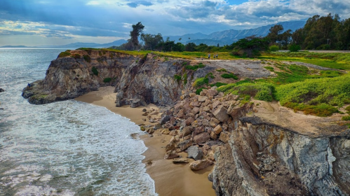 A scenic coastal view featuring rocky cliffs, sandy beach, and waves under a cloudy sky with distant mountains.