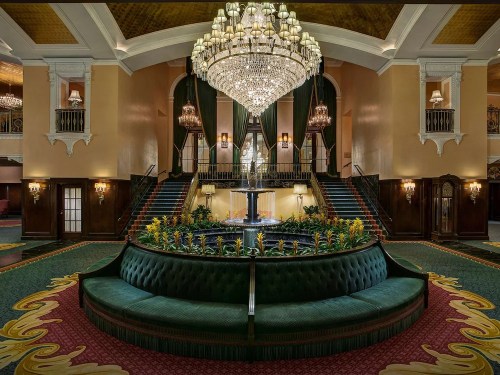 Elegant hotel lobby featuring a grand chandelier, lush plants, and a circular green seating area around a fountain.