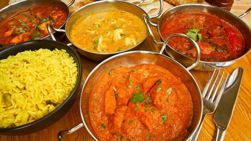 A variety of colorful Indian dishes in metal bowls, including curries and yellow rice, on a wooden table.