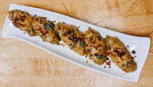 A white platter with five spicy, seasoned chicken wings garnished with herbs and red pepper flakes on a wooden surface.
