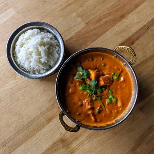A bowl of creamy curry with peas and diced vegetables, served alongside a small bowl of white rice on a wooden surface.
