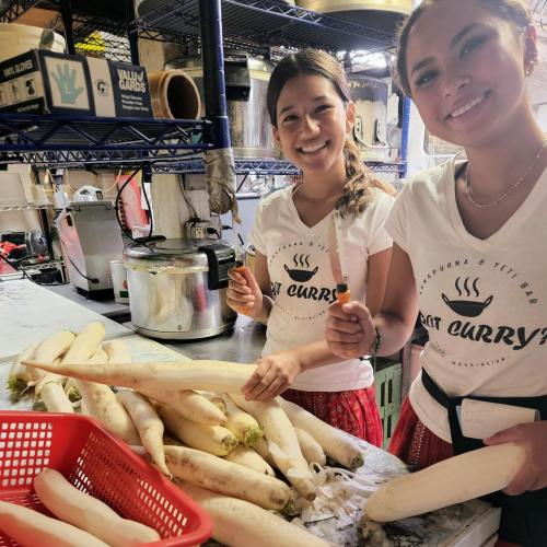 Two smiling women in a kitchen, preparing large white radishes on a countertop.