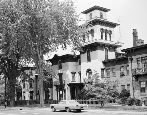 Historic building with a tall tower, surrounded by trees, and a vintage car parked in front. Black and white photo.