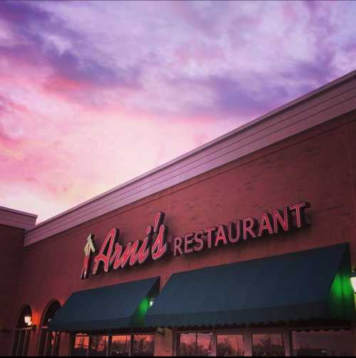 Exterior of Arni's Restaurant with a colorful sunset sky in the background. Neon sign illuminated.