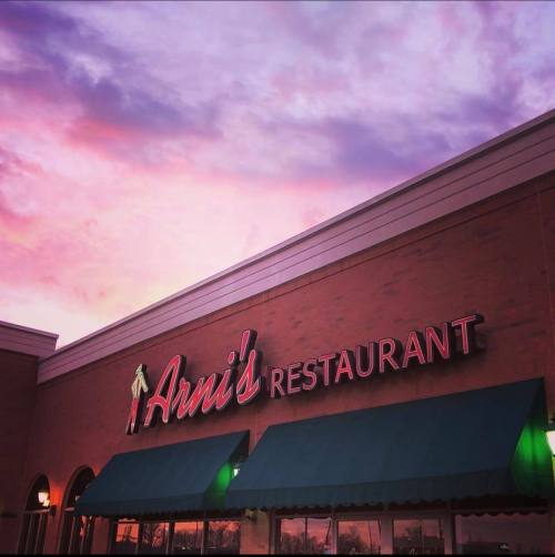 Exterior of Arni's Restaurant with a colorful sunset sky in the background. Neon sign illuminated.