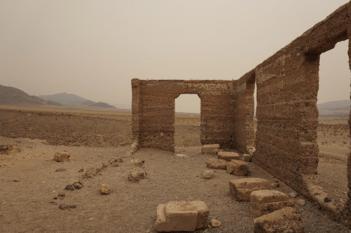 Ruins of a stone structure in a barren landscape, with mountains in the background and a hazy sky.