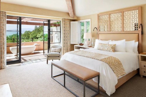 Bright, modern bedroom with a large bed, seating area, and balcony overlooking greenery. Natural light fills the space.
