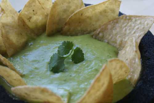 A bowl of green dip surrounded by crispy tortilla chips, garnished with a sprig of cilantro.