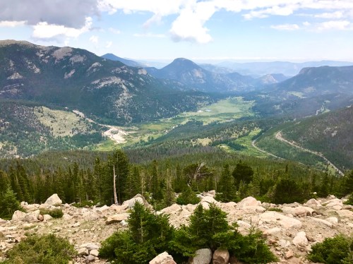 A panoramic view of a lush valley surrounded by mountains, with a winding river and scattered clouds in the sky.