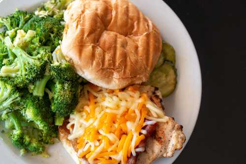 A plate with grilled chicken topped with cheese, a bun, broccoli, and pickles.