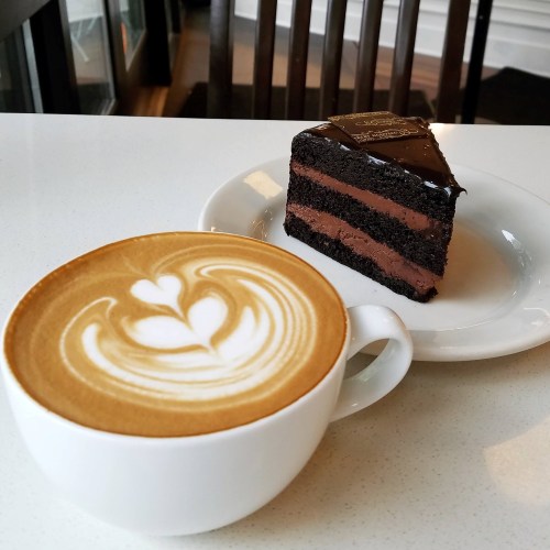 A cup of latte with heart-shaped foam next to a slice of chocolate layer cake on a white plate.