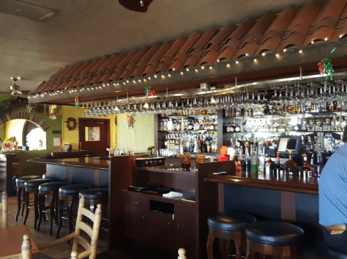 A cozy restaurant bar with a tiled roof, shelves of drinks, and festive decorations. Bar stools line the counter.