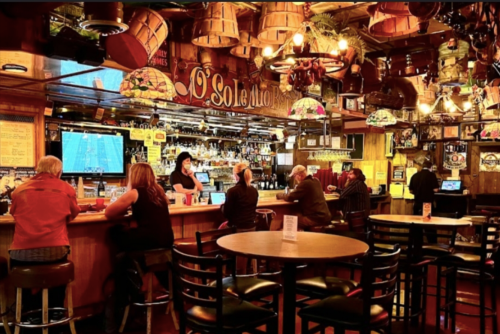 A cozy bar scene with patrons seated at tables and the bar, colorful lights, and a lively atmosphere.