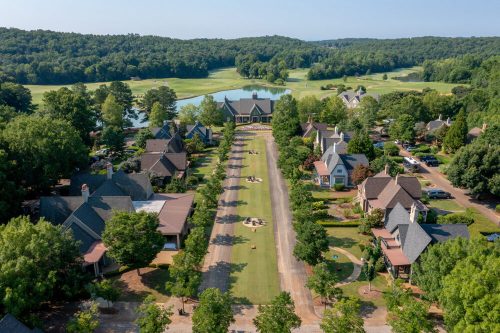 Aerial view of a scenic neighborhood with homes, a green lawn, and a lake surrounded by trees.