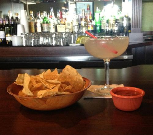 A bowl of tortilla chips with salsa and a margarita on a bar counter, with a blurred liquor shelf in the background.