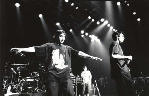 Two performers on stage in black shirts, with a band setup and bright stage lights in the background.