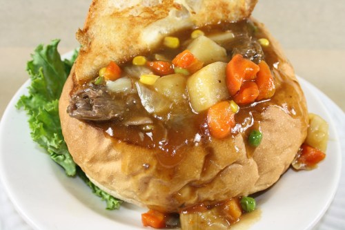 A hearty bread bowl filled with beef stew, vegetables, and gravy, served on a plate with a lettuce leaf.