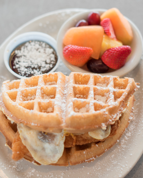 A plate featuring a waffle sandwich with fried chicken, powdered sugar, and a side of fresh fruit and syrup.