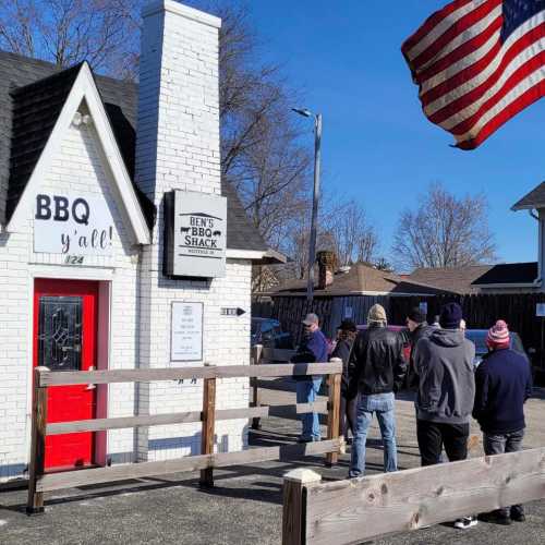 A group of people waiting in line outside Ben's BBQ Shack, with an American flag waving in the background.