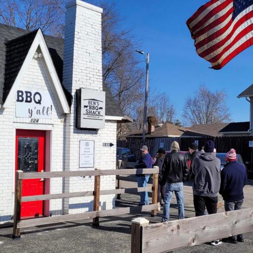 A group of people waiting in line outside Ben's BBQ Shack, with an American flag waving in the background.