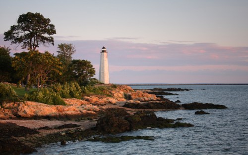 A lighthouse stands on a rocky shore at sunset, surrounded by trees and calm waters.