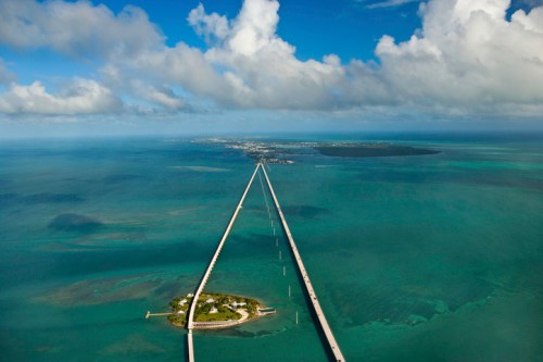Aerial view of a long bridge extending over turquoise waters, leading to a small island under a partly cloudy sky.