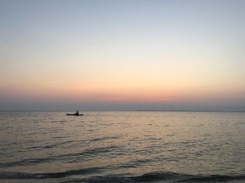 A serene sunset over calm waters, with a lone fisherman in a small boat silhouetted against the horizon.