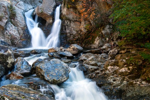A serene waterfall cascades over rocks, surrounded by lush greenery and autumn foliage.