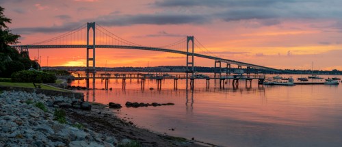 A sunset view of a bridge over water, with boats docked and rocky shoreline in the foreground.
