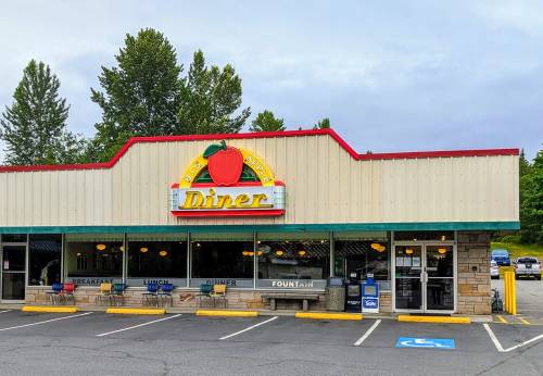 A colorful diner with a large apple sign, featuring outdoor seating and a parking lot surrounded by trees.