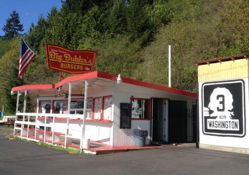 A roadside burger stand with a red and white exterior, featuring a large sign and an American flag. Surrounded by trees.