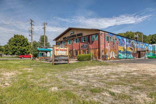 A colorful BBQ restaurant with a mural, surrounded by grass and trees, under a clear blue sky.