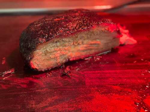 A close-up of a smoked brisket, showcasing its dark, charred exterior and tender, juicy interior on a wooden surface.