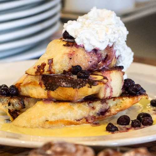Stack of French toast topped with blueberries and whipped cream, drizzled with syrup on a white plate.