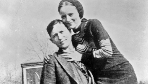 A vintage black-and-white photo of a man and woman posing together, smiling, outdoors with trees in the background.