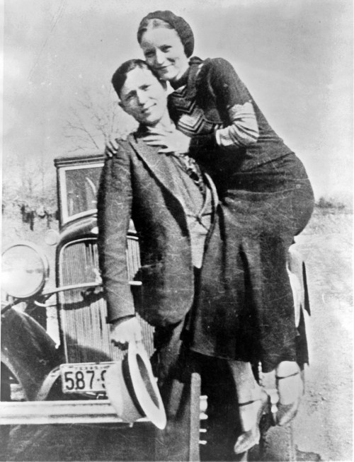 A vintage black-and-white photo of a man and woman posing together on a car, smiling at the camera.