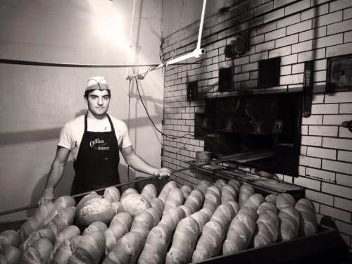 A baker stands in front of a large oven, surrounded by freshly baked loaves of bread in a vintage setting.