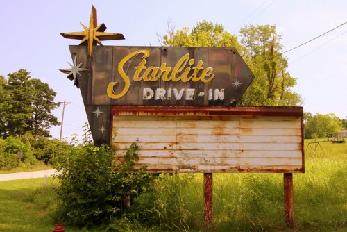 Rusty vintage sign for "Starlite Drive-In" with a star design, surrounded by overgrown grass and trees.