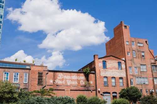 A brick building with faded signage under a blue sky with fluffy clouds, surrounded by greenery.