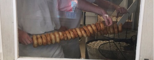 A person holds a long skewer of freshly fried donuts, with a fryer visible in the background.