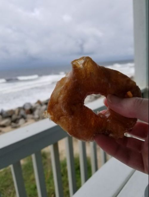 A hand holding a glazed donut with a beach and ocean in the background under a cloudy sky.