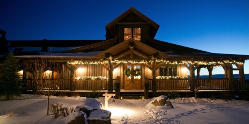 A cozy log cabin decorated with lights, surrounded by snow, at dusk with a clear blue sky.