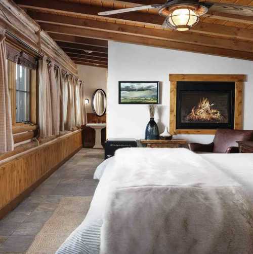 Cozy bedroom with a fireplace, wooden accents, and large windows, featuring a bed with a soft blanket and a chair.