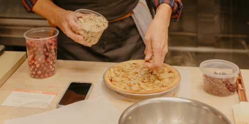 A person sprinkles topping on an apple pie, with containers of berries and a phone nearby on a kitchen counter.