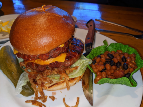 A hearty burger with cheese, lettuce, and crispy onions, served with beans and a pickle on the side.