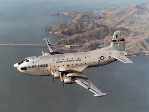 A U.S. Air Force military transport aircraft flying over a coastal landscape with mountains and water below.